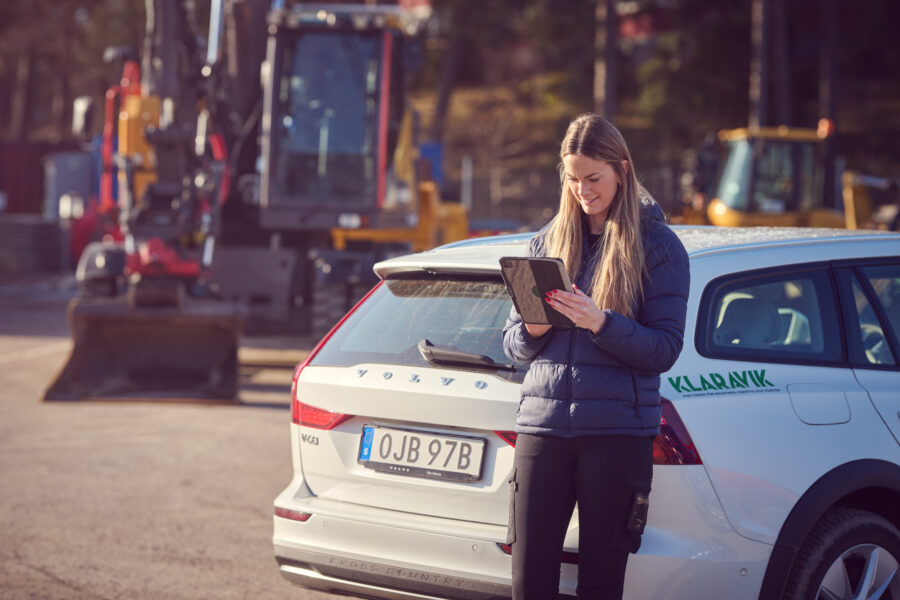 Auktionsm&auml;klare fr&aring;n Klaravik st&aring;r vid en Klaravik-bil och uppdaterar auktionsunderlag p&aring; en surfplatta.